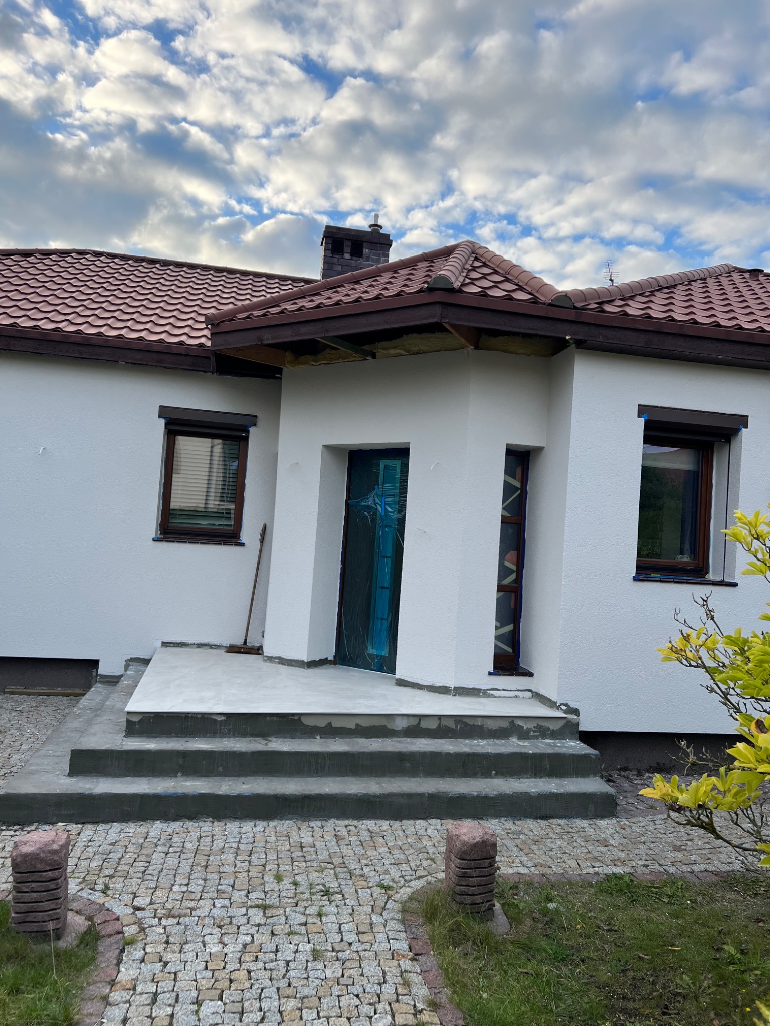 Biały dom z brązowym dachem w trakcie wykończenia elewacji i schodów wejściowych, zabezpieczone okna i drzwi folią, widoczna kostka brukowa przed domem.