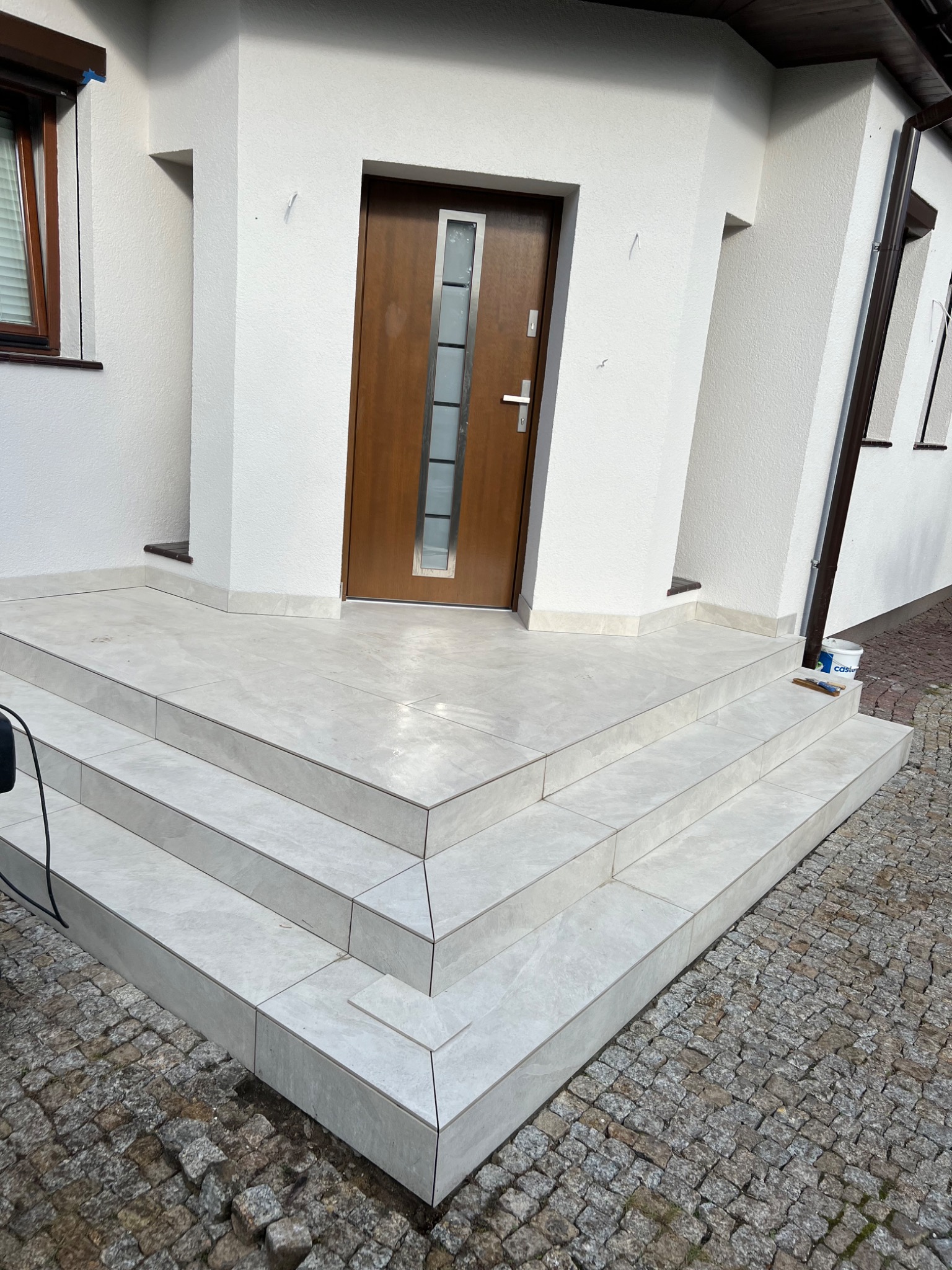 Wykończone płytkami schody wejściowe do domu z brązowymi drzwiami i jasną elewacją, widoczny fragment kostki brukowej i wiaderko po farbie.