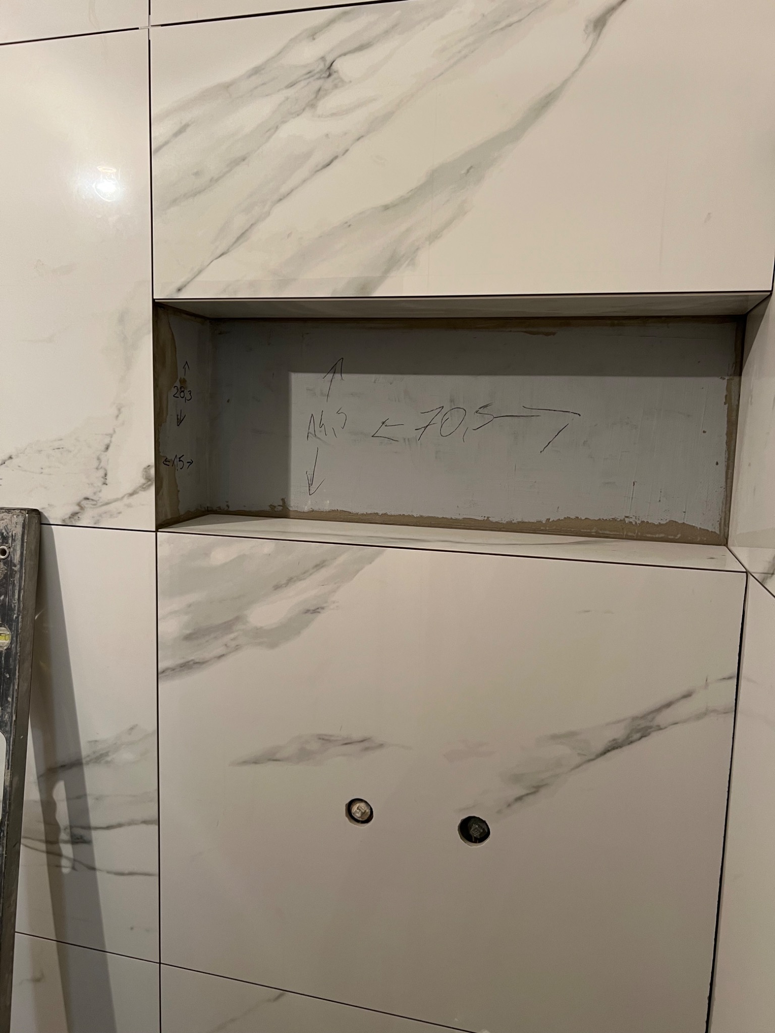 Ściana z białych płytek imitujących marmur z wnęką, wewnątrz której widoczne są wymiary naniesione ołówkiem, poniżej dwa otwory na rury.