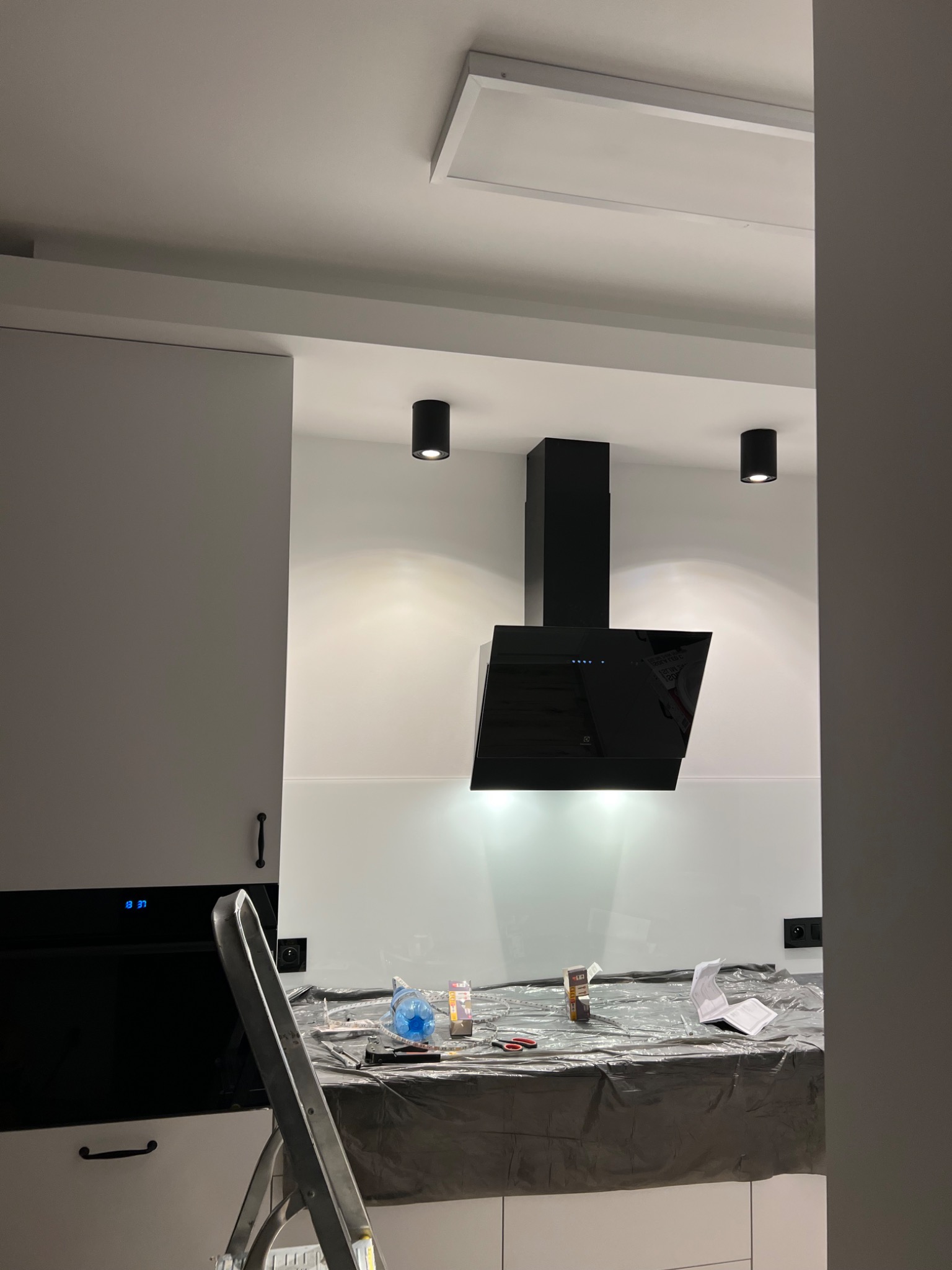 Nowoczesna kuchnia w trakcie montażu okapu: czarny okap kominowy na tle białych płytek, zabezpieczony blat z widoczną drabiną i narzędziami.