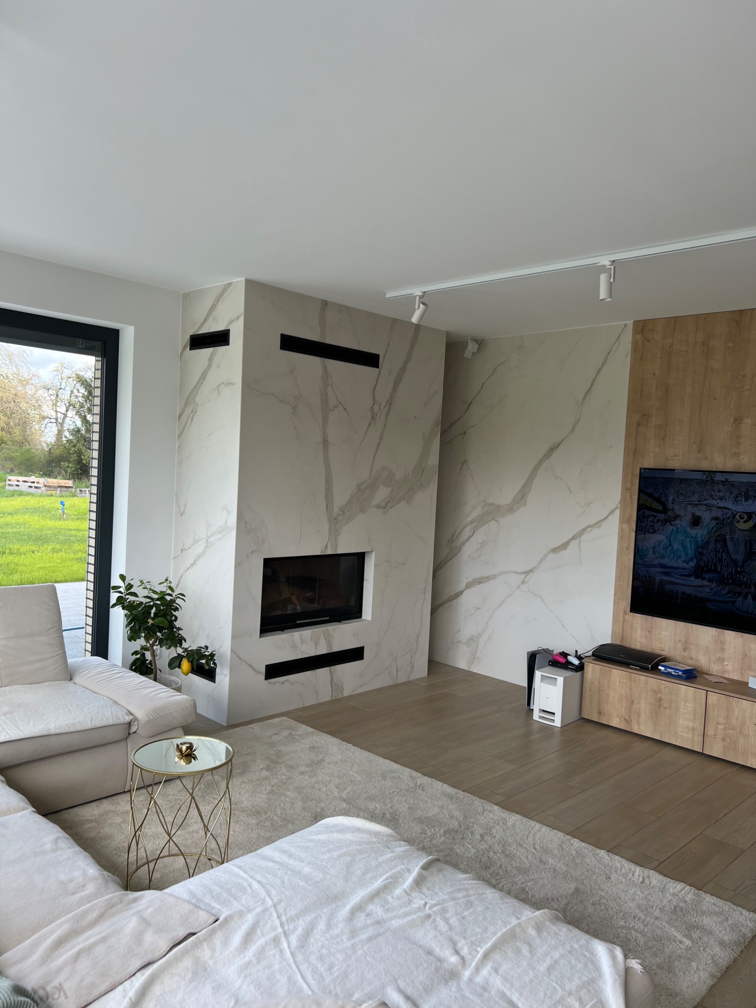 Nowoczesny salon z marmurowym kominkiem i drewnianą ścianą telewizyjną, minimalistyczny design, jasne wnętrze z widokiem na ogród przez duże okno.