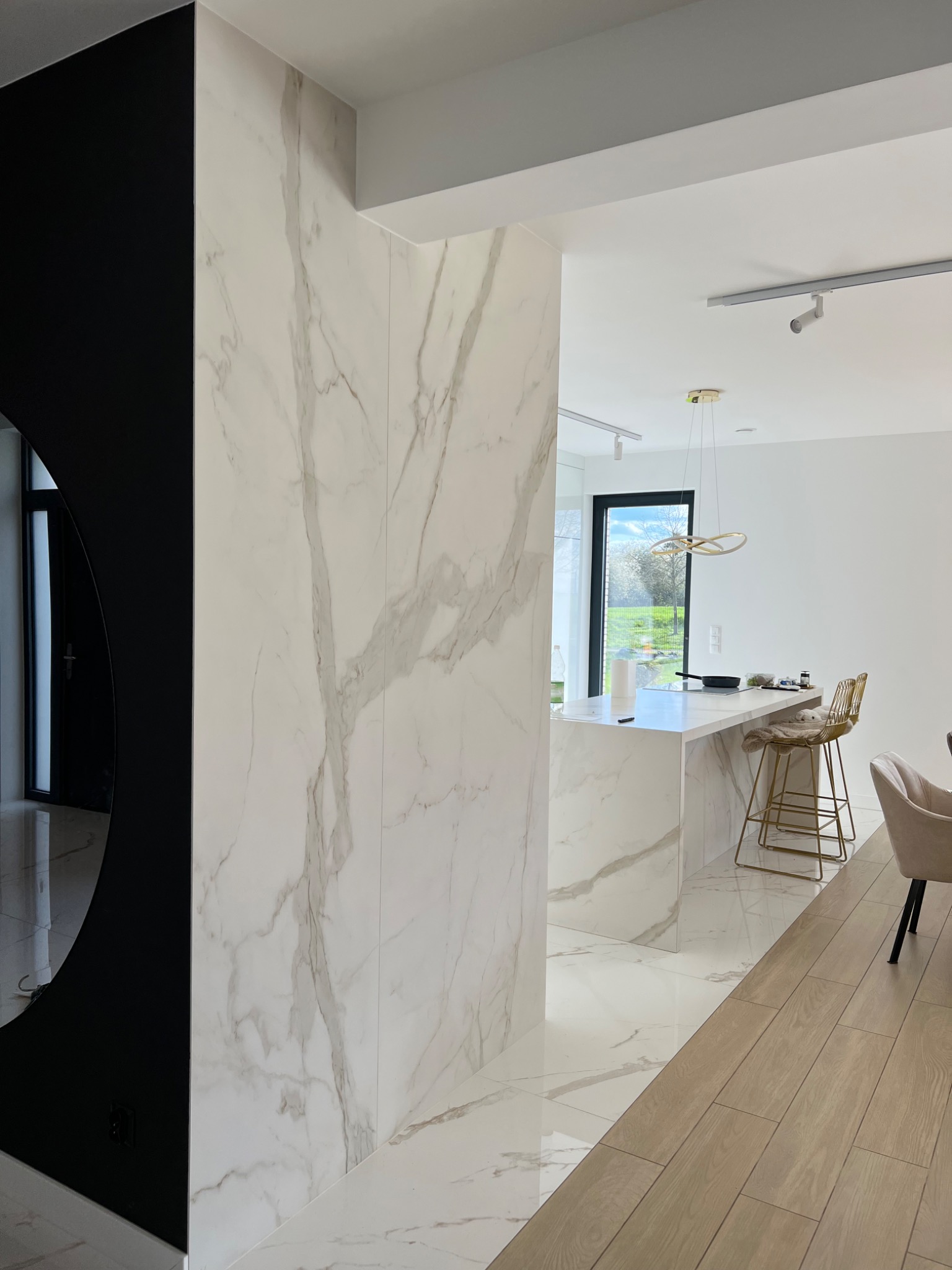 Nowoczesne wnętrze z marmurowymi płytami na ścianach i podłodze, widoczna kuchnia z wyspą i złotymi hokerami, czarny element dekoracyjny z lustrem, drewniana podłoga w części jadalnej.