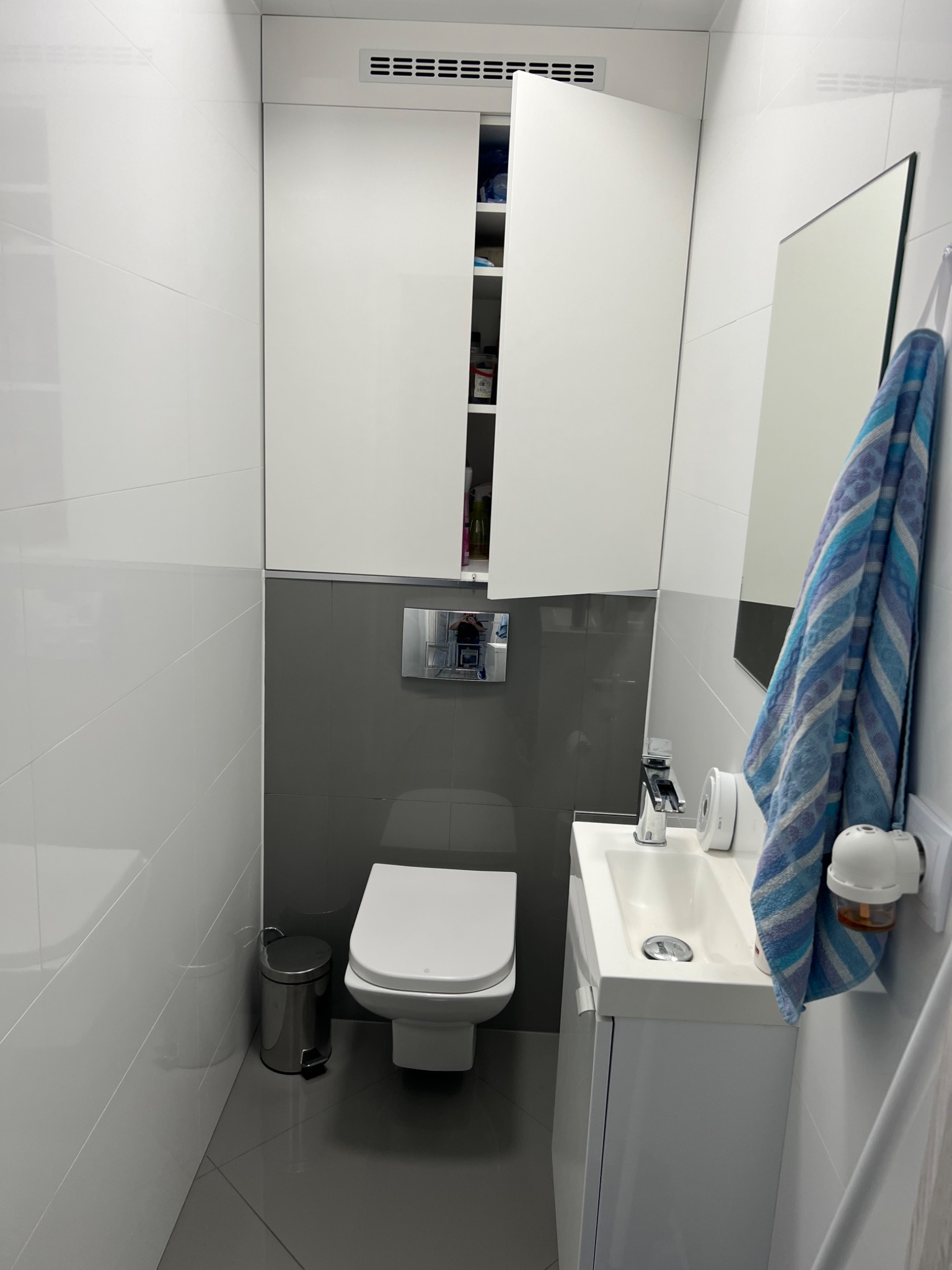 Nowoczesna, mała łazienka z białymi płytkami i szarą strefą przy toalecie, wisząca szafka z otwartymi drzwiczkami, biała umywalka z szafką, lustro i niebiesko-biały ręcznik.