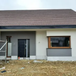 Ocieplona elewacja domu jednorodzinnego w trakcie realizacji, widoczne rusztowania przy oknie, szara i biała kolorystyka ścian, brązowa dachówka.