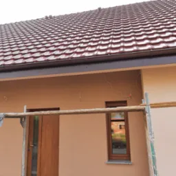 Elewacja budynku jednorodzinnego w trakcie prac wykończeniowych, widoczne rusztowanie, brązowe okna i drzwi, oraz dachówka w odcieniach czerwieni i brązu.