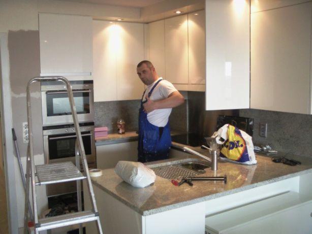 Montaż białych mebli kuchennych w Bielsku Podlaskim. Widoczny instalator w trakcie pracy oraz drabina. Nowoczesny design i oświetlenie podkreślają estetykę wnętrza.