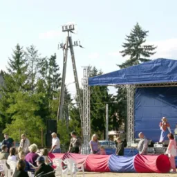 Plenerowa scena z niebieskim zadaszeniem, grupa dzieci na scenie z mikrofonami, publiczność siedząca na krzesłach, widok na dom z czerwonym dachem w tle.