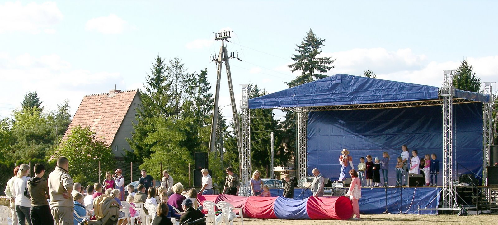 Plenerowa scena z niebieskim zadaszeniem, grupa dzieci na scenie z mikrofonami, publiczność siedząca na krzesłach, widok na dom z czerwonym dachem w tle.