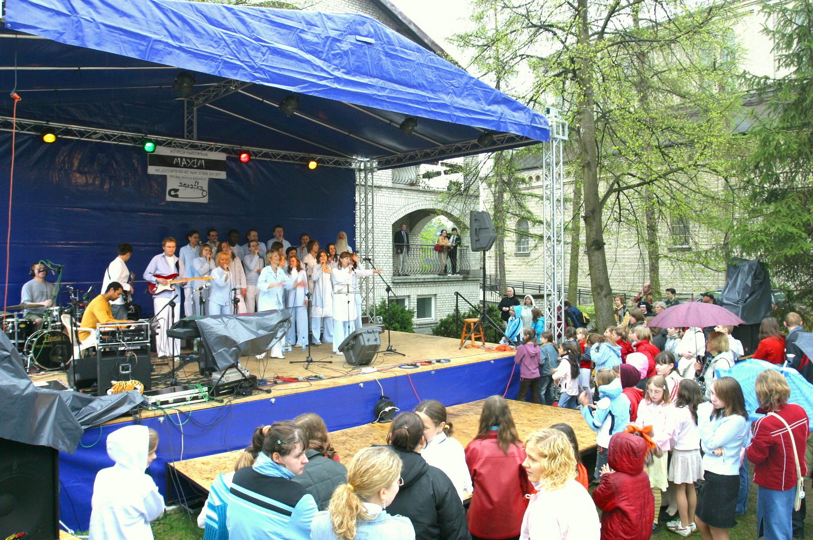 Chór w jasnych strojach występuje na scenie pod niebieskim zadaszeniem, nagłośnienie widoczne, publiczność obserwuje występ na tle budynku.