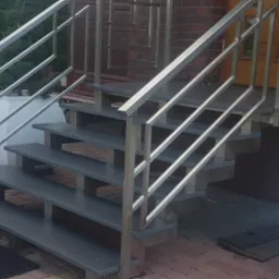 Zewnętrzne schody z szarymi stopniami i nowoczesną, prostą balustradą ze stali nierdzewnej, prowadzące do drzwi wejściowych domu z cegły.