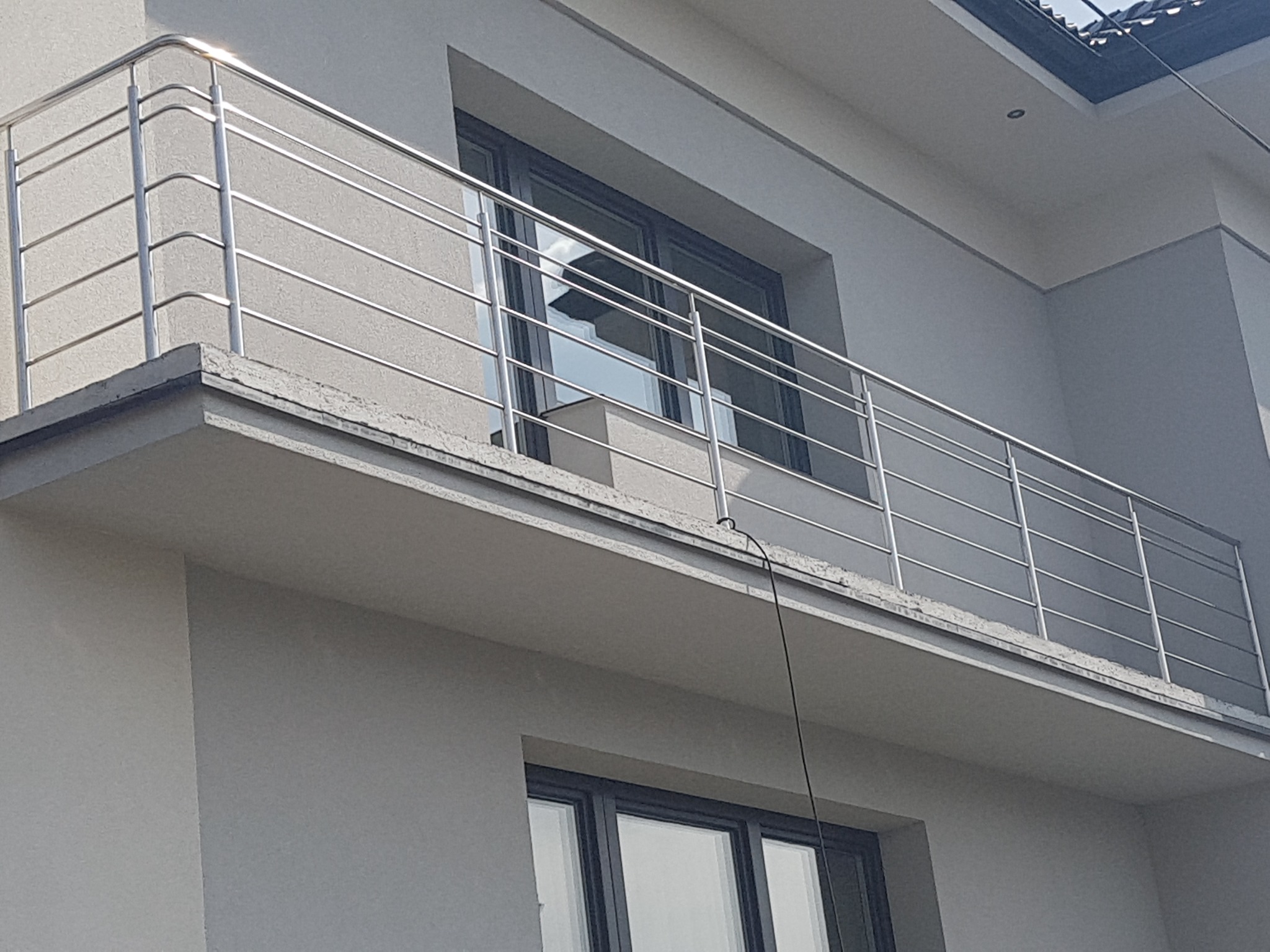 Balkon z nowoczesną, metalową balustradą o poziomym układzie prętów, zamontowaną na szarym budynku mieszkalnym z widocznymi oknami.