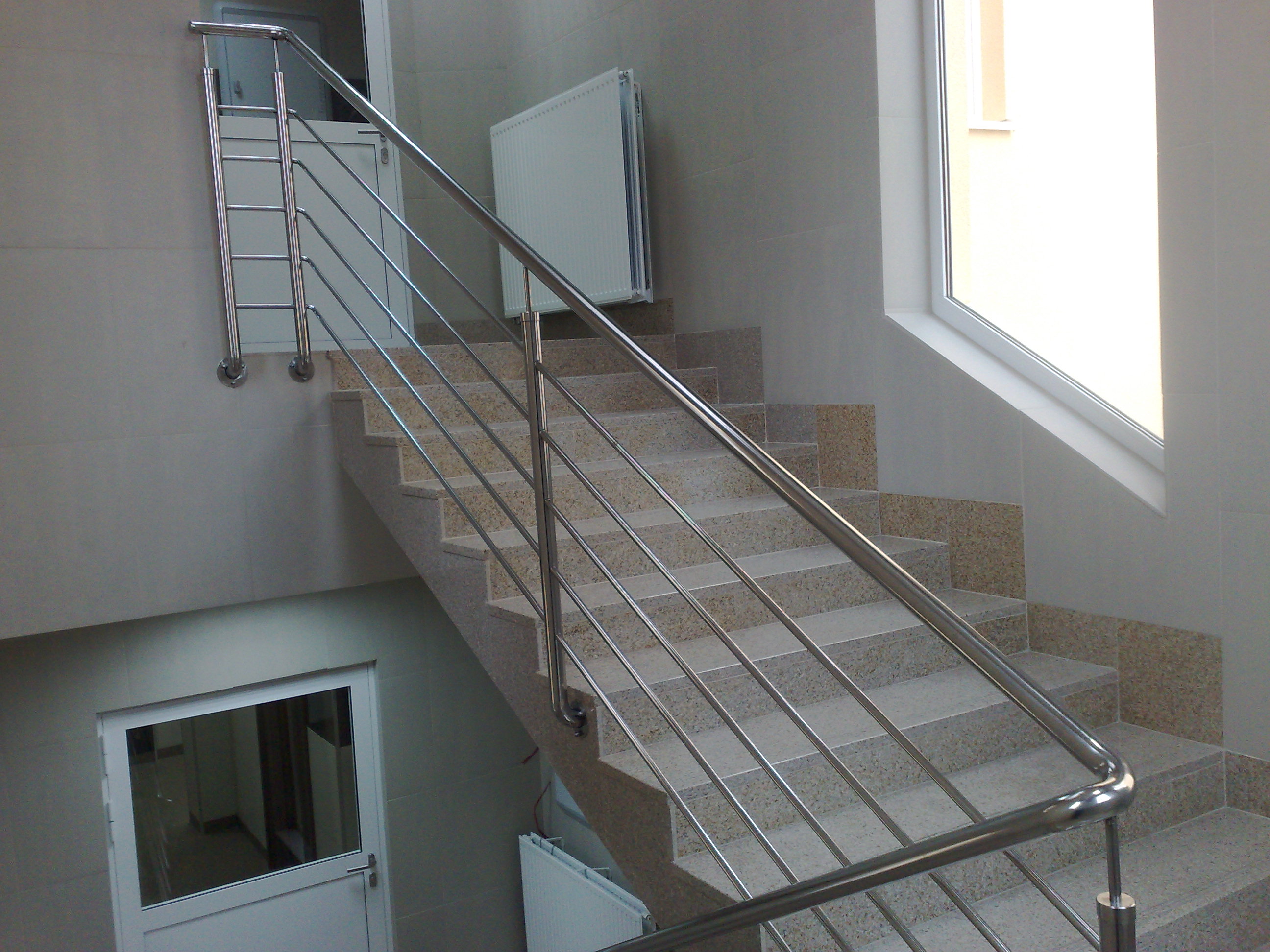 Wewnętrzne schody z kamiennymi stopniami i poręczą ze stali nierdzewnej o minimalistycznym designie, widoczne białe drzwi i grzejnik.