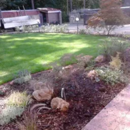 Ukończony ogród z trawnikiem, rabatami wysypanymi korą i kamieniami. Widoczny fragment chodnika i zaparkowany samochód dostawczy.
