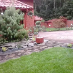 Ogród z trawnikiem i ścieżką z kamieni prowadzącą do budynku z czerwoną dachówką. Widoczne ozdobne drzewa i krzewy, całość tworzy harmonijny krajobraz.