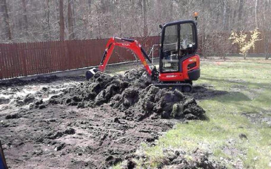 Czerwona mini koparka Kubota na działce z rozkopaną ziemią, widoczny fragment drewnianego ogrodzenia i drzewa w tle, słoneczny dzień.