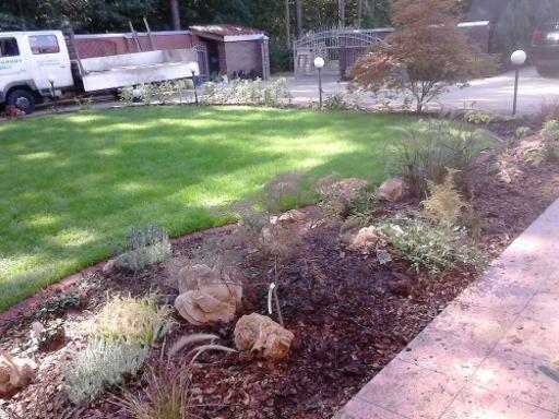 Ukończony ogród z trawnikiem, rabatami wysypanymi korą i kamieniami. Widoczny fragment chodnika i zaparkowany samochód dostawczy.
