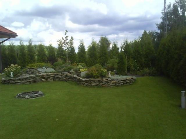 Aranżacja ogrodu z kamiennym murkiem, rabatami kwiatowymi i równo przystrzyżonym trawnikiem na tle iglaków i pochmurnego nieba. Widok na zagospodarowaną przestrzeń.