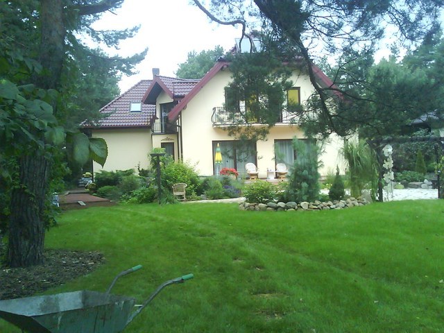 Rozległy, zadbany trawnik przed domem z beżową elewacją i czerwoną dachówką, z widocznym w tle fragmentem oczka wodnego i dekoracyjnymi nasadzeniami. W pierwszym planie, lekko po lewej, widoczna...