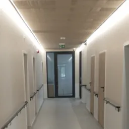 Długi korytarz w budynku użyteczności publicznej z szarymi ścianami, poręczami i drzwiami na końcach, oświetlony liniowymi lampami LED pod sufitem z perforowanej blachy.