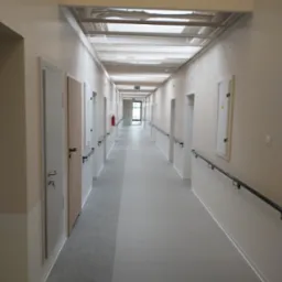 Długi korytarz w budynku użyteczności publicznej z wieloma drzwiami, poręczą i kratką wentylacyjną pod sufitem, widoczny znak ewakuacyjny.