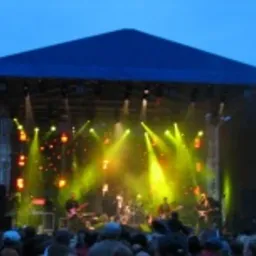 Scena plenerowa z oświetleniem estradowym i zespołem muzycznym podczas występu. Widoczne reflektory, konstrukcja sceniczna z niebieskim zadaszeniem i sylwetki muzyków.