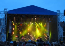 Scena plenerowa z oświetleniem estradowym i zespołem muzycznym podczas występu. Widoczne reflektory, konstrukcja sceniczna z niebieskim zadaszeniem i sylwetki muzyków.