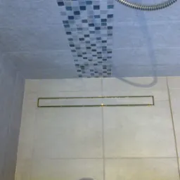 Ułożona jasnoszara glazura w kabinie prysznicowej z odpływem liniowym i dekoracyjnym pasem mozaiki na ścianie.