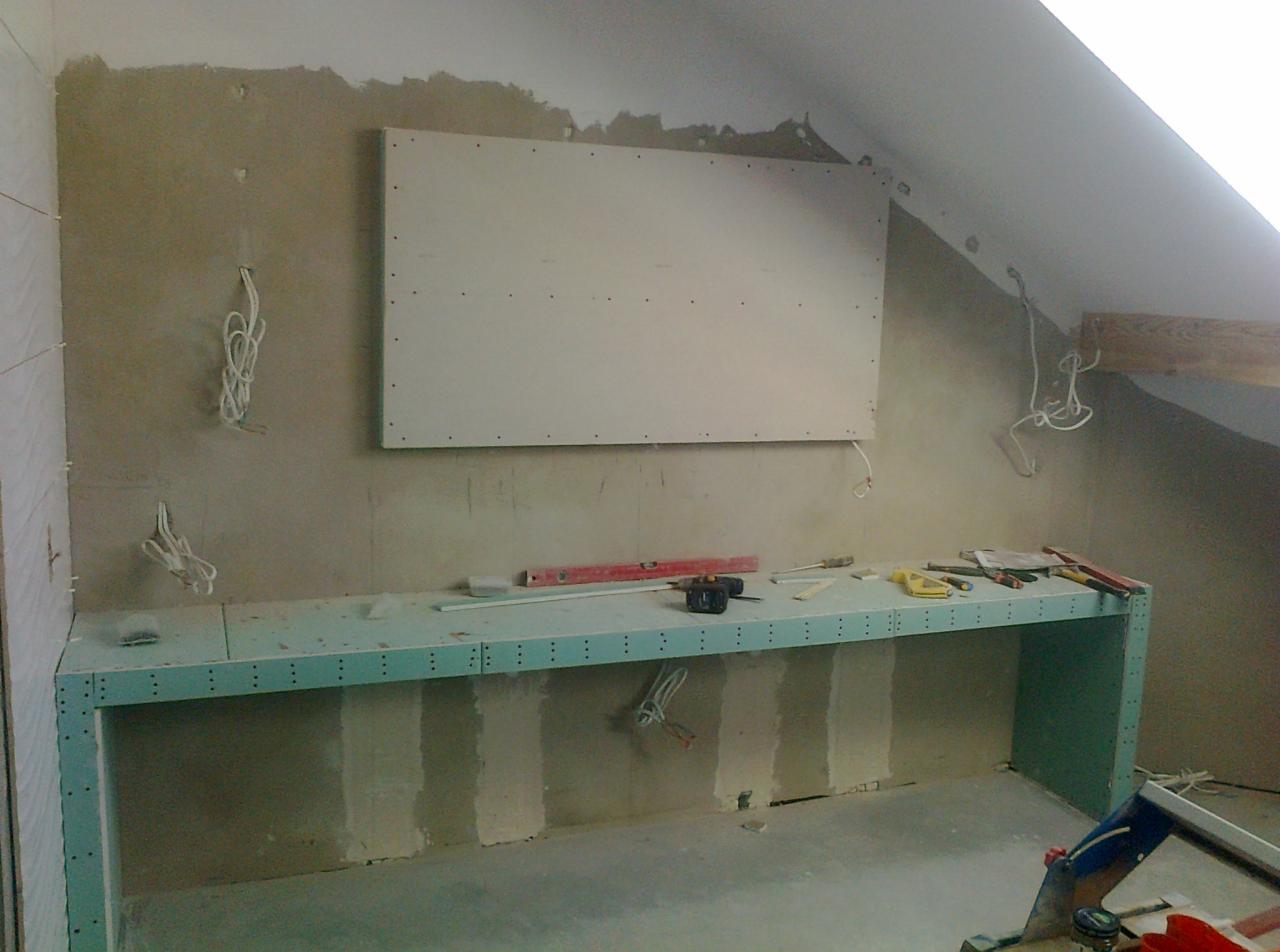 Poddasze w trakcie prac wykończeniowych: płyta gipsowo-kartonowa zamontowana na ścianie, widoczne kable elektryczne, blat roboczy z narzędziami, ściany w stanie surowym.