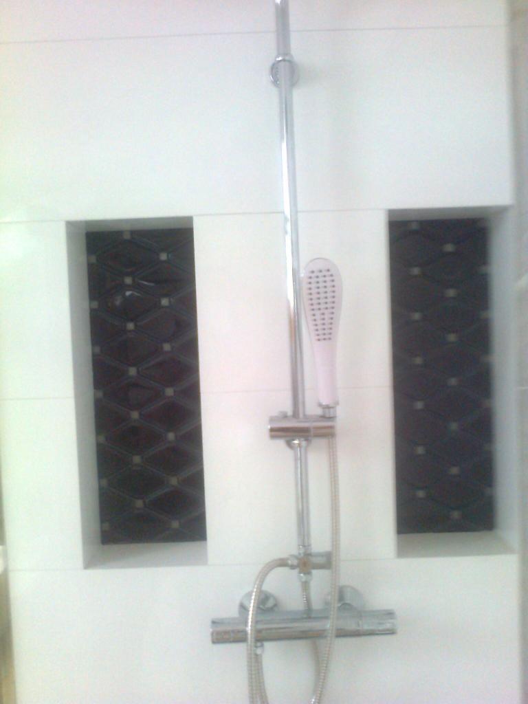 Biała ściana w łazience z wnękami wyłożonymi ciemną, wzorzystą glazurą i zamontowaną chromowaną baterią prysznicową z białą słuchawką.