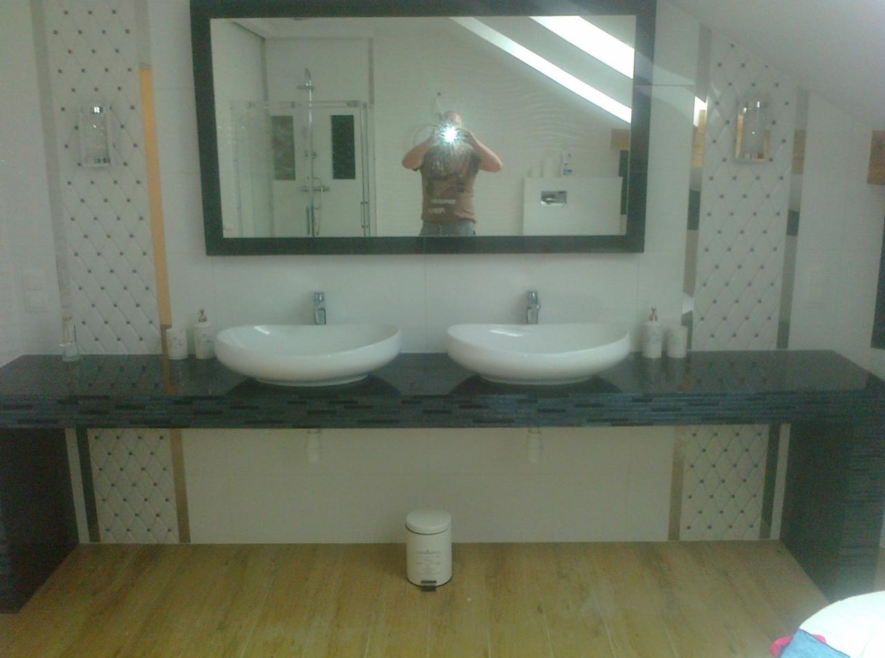 Dwie białe, owalne umywalki nablatowe na ciemnym blacie z mozaiką, duże lustro z czarną ramą odbijające część łazienki, białe płytki imitujące pikowanie na ścianach, drewnopodobna podłoga.