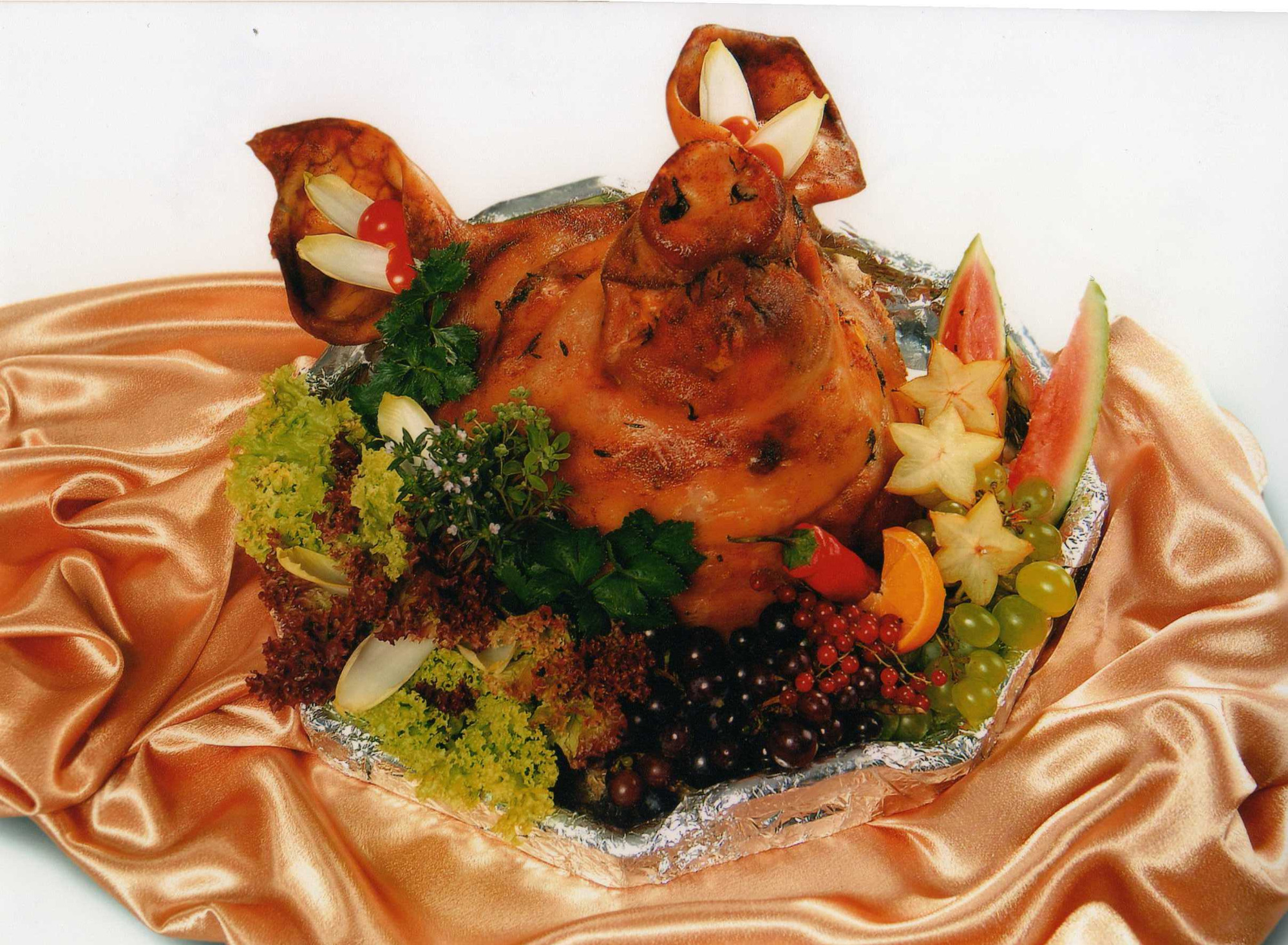 Pieczone prosię podane na srebrnej tacy, udekorowane sałatą, winogronami, karambolą, arbuzem i innymi owocami, leżące na pomarańczowej, satynowej tkaninie.