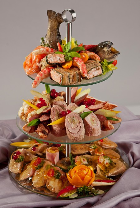 Trzypoziomowa patera z różnorodnymi przekąskami: wędzony łosoś, roladki mięsne, galaretki rybne i dekoracje z warzyw na tle fioletowego materiału.