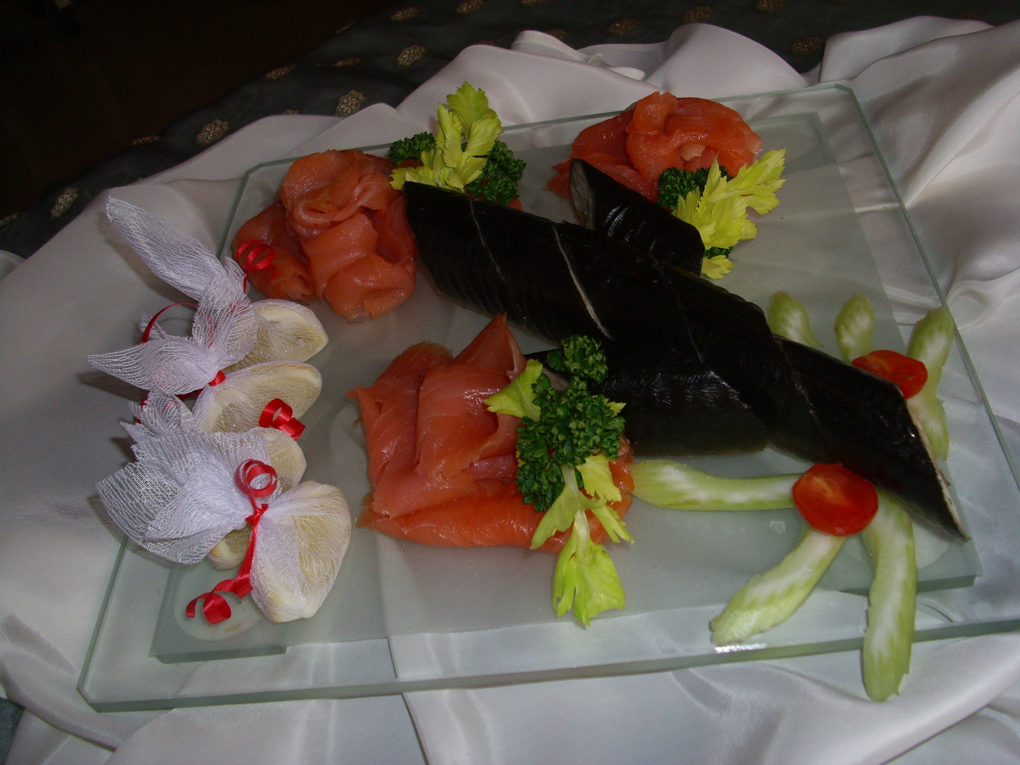 Elegancko podane przystawki na szklanym talerzu: roladki z łososia, ciemne rulony rybne, seler naciowy z pomidorkami koktajlowymi i woreczki z jasnymi kulkami zawiązane czerwoną wstążką.
