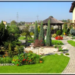 Pełen detali ogród z oczkiem wodnym, ścieżką z kostki, altaną z trzcinowym dachem i różnorodną roślinnością, w tym strzyżonymi drzewami i barwnymi kwiatami, na tle zabudowań.