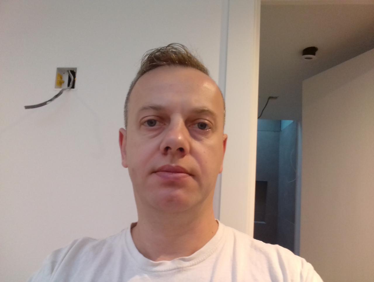 Mężczyzna na tle ściany z wystającymi kablami elektrycznymi i widokiem na wnętrze pomieszczenia w trakcie remontu.