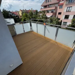 Nowoczesny balkon z brązową deską kompozytową, szklane balustrady i widok na budynki mieszkalne w słoneczny dzień. Wykończony taras.