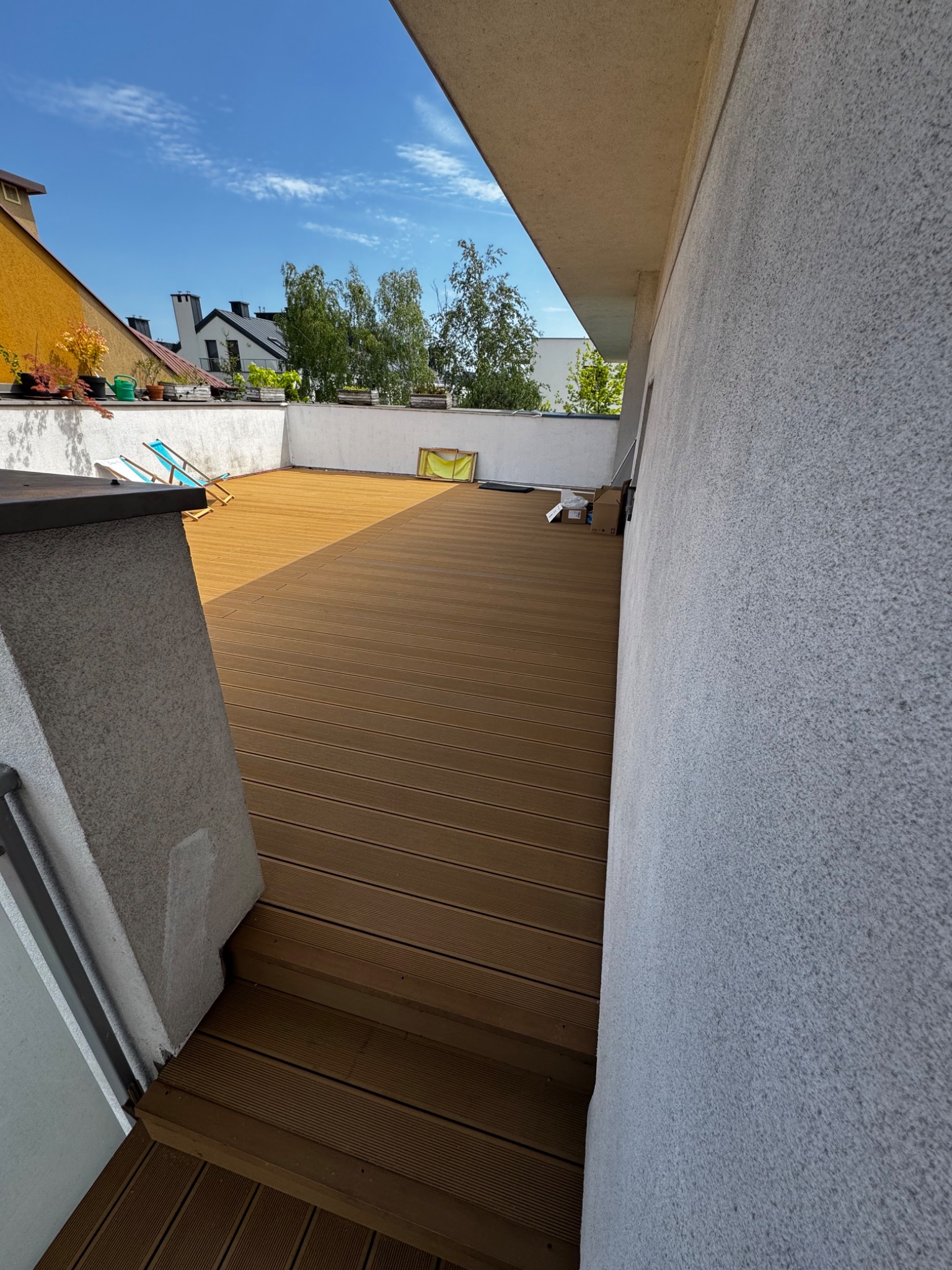 Przestronny taras na dachu z brązową podłogą, widok z góry, niebieskie leżaki i skrzynki na rośliny w tle, budynki, błękitne niebo.