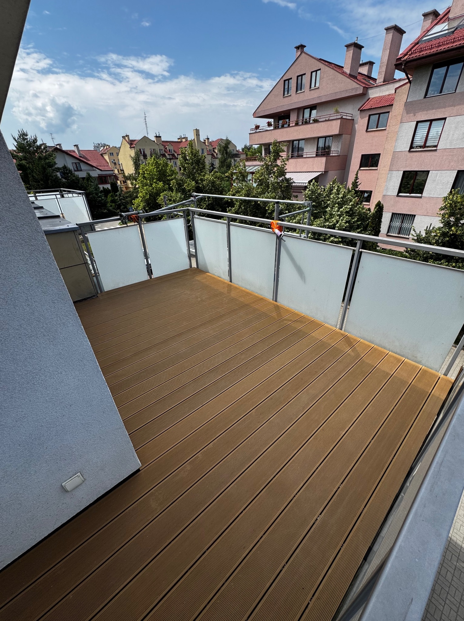 Nowoczesny balkon z brązową deską kompozytową, szklane balustrady i widok na budynki mieszkalne w słoneczny dzień. Wykończony taras.