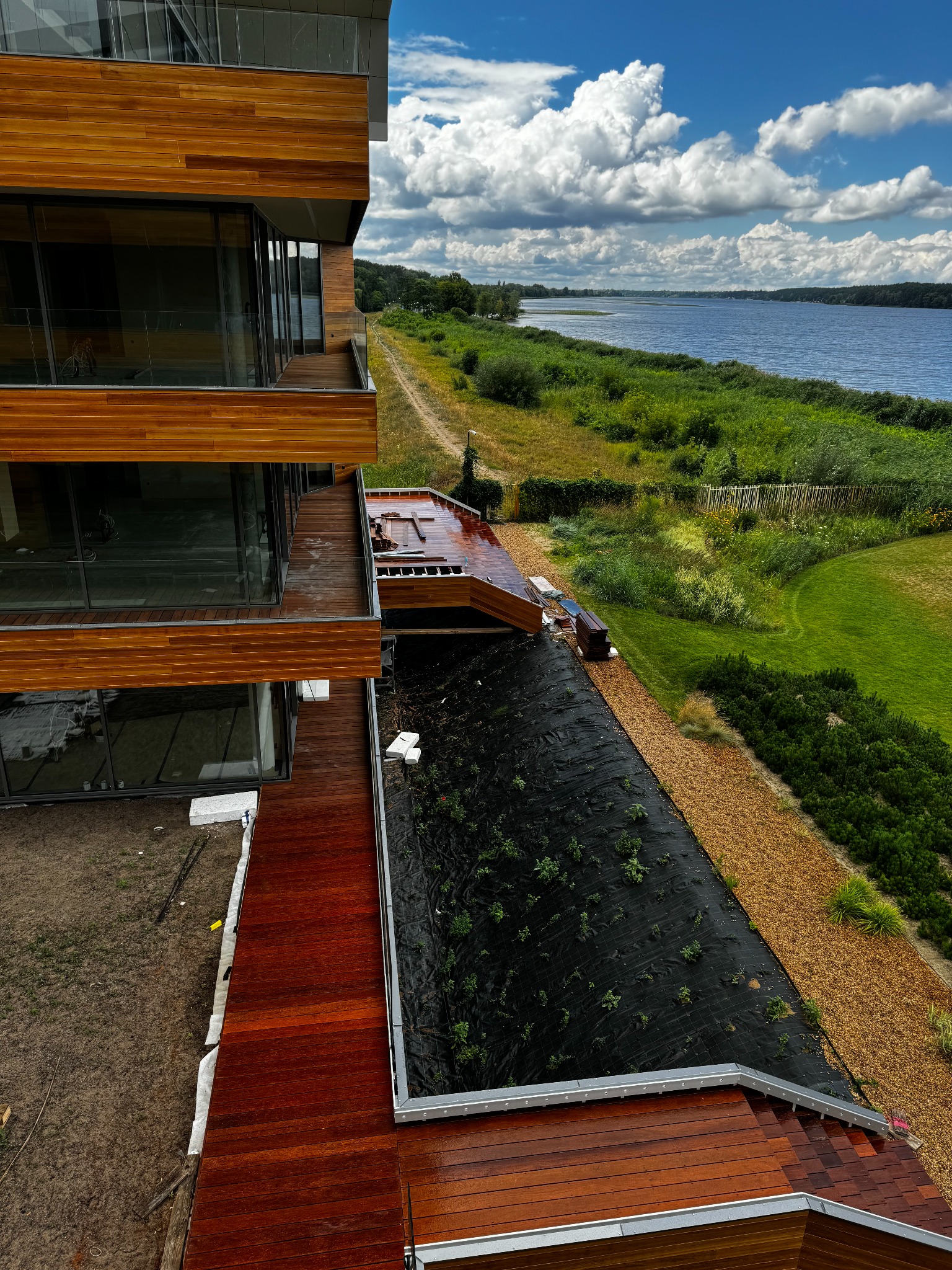 Nowoczesny budynek z tarasami z egzotycznego drewna, widok z góry na etap budowy, z widokiem na jezioro i błękitne niebo z chmurami.