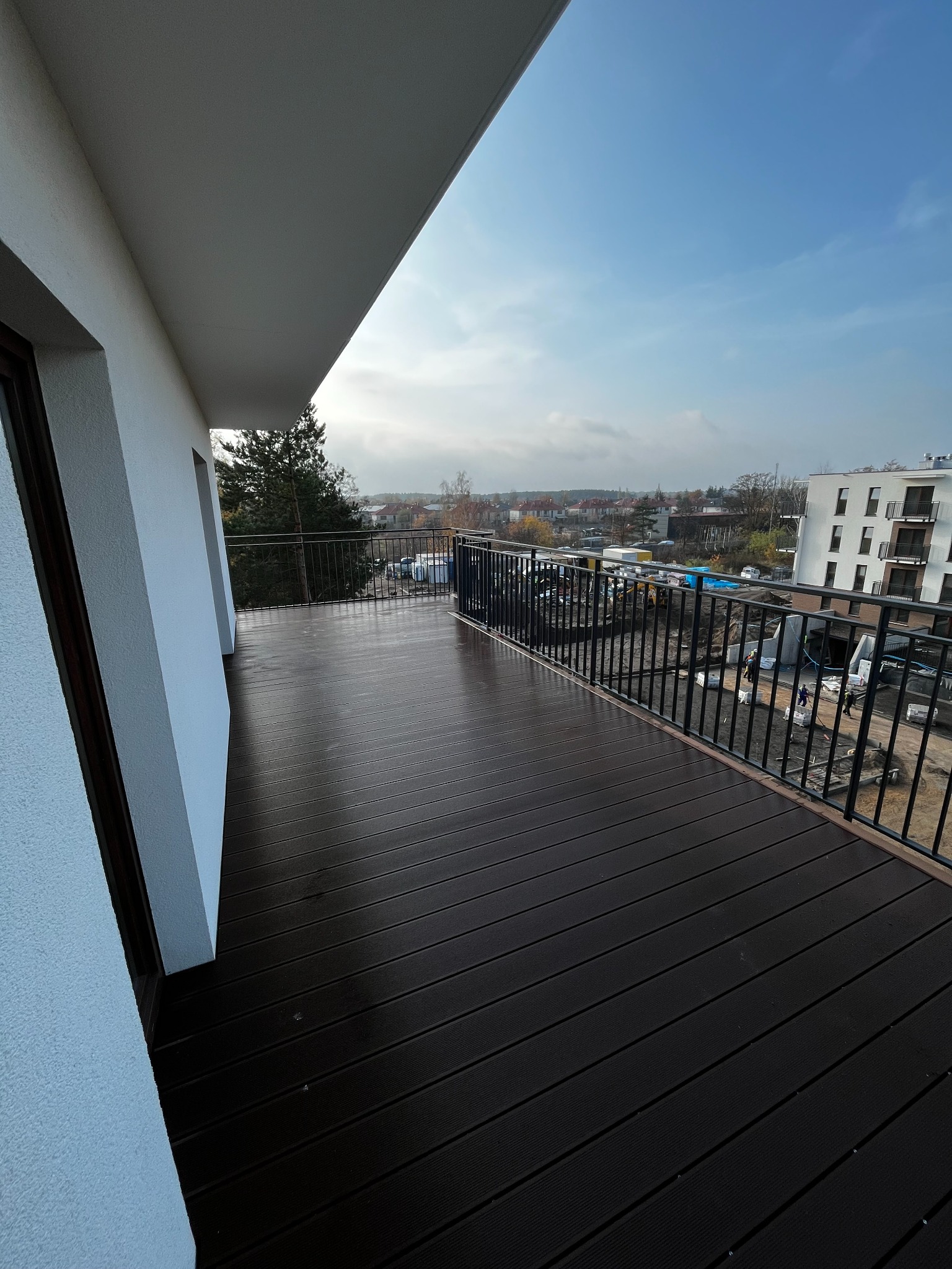 Nowoczesny balkon z ciemną podłogą i czarną balustradą, widok na miasto w tle. Minimalistyczny design, jasne ściany, świeże powietrze. Perspektywa z narożnika balkonu.