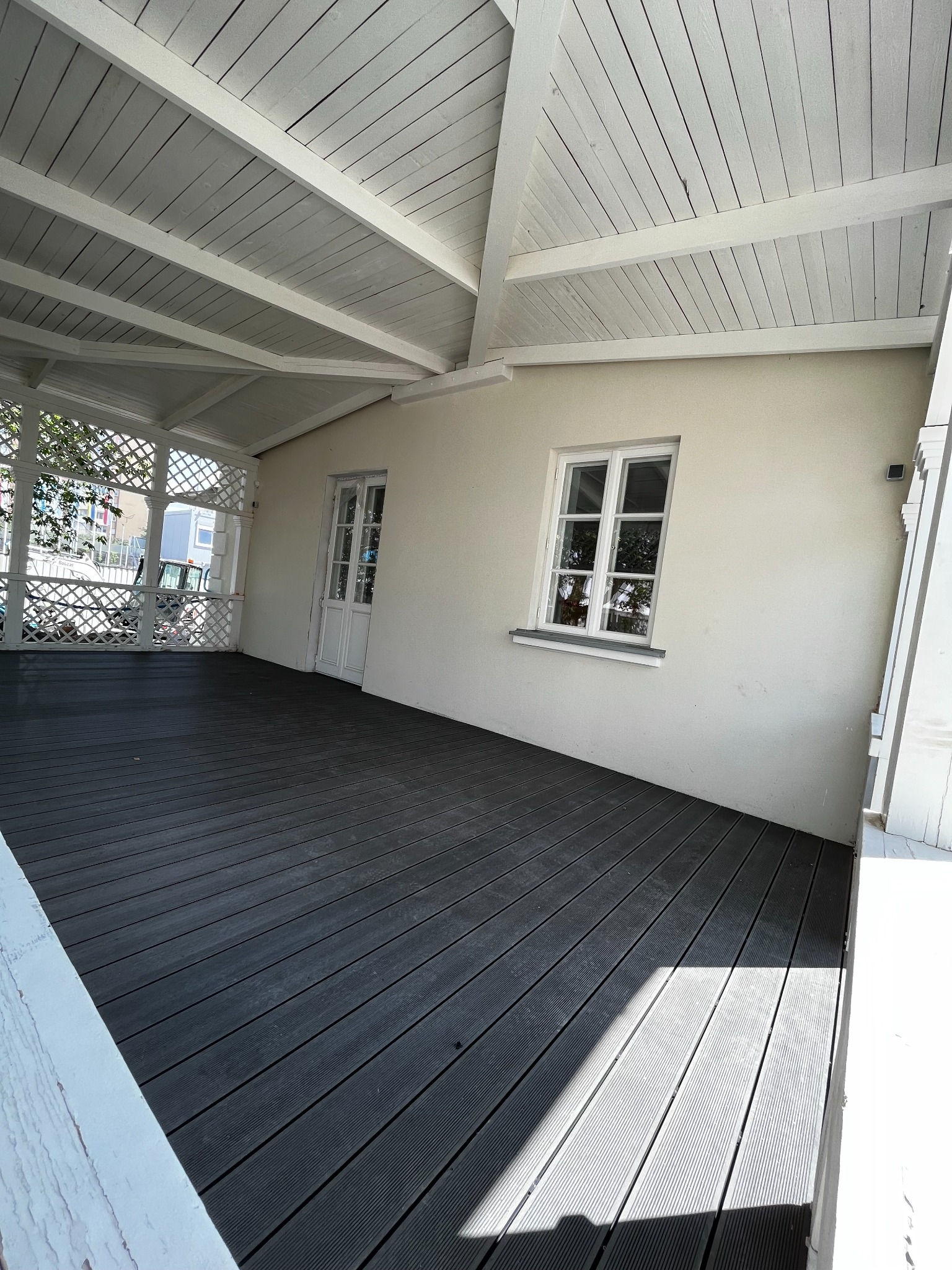 Zadaszony taras z szarą podłogą, białą konstrukcją i ażurową balustradą. Widoczne okno i drzwi. Jasne, naturalne światło.