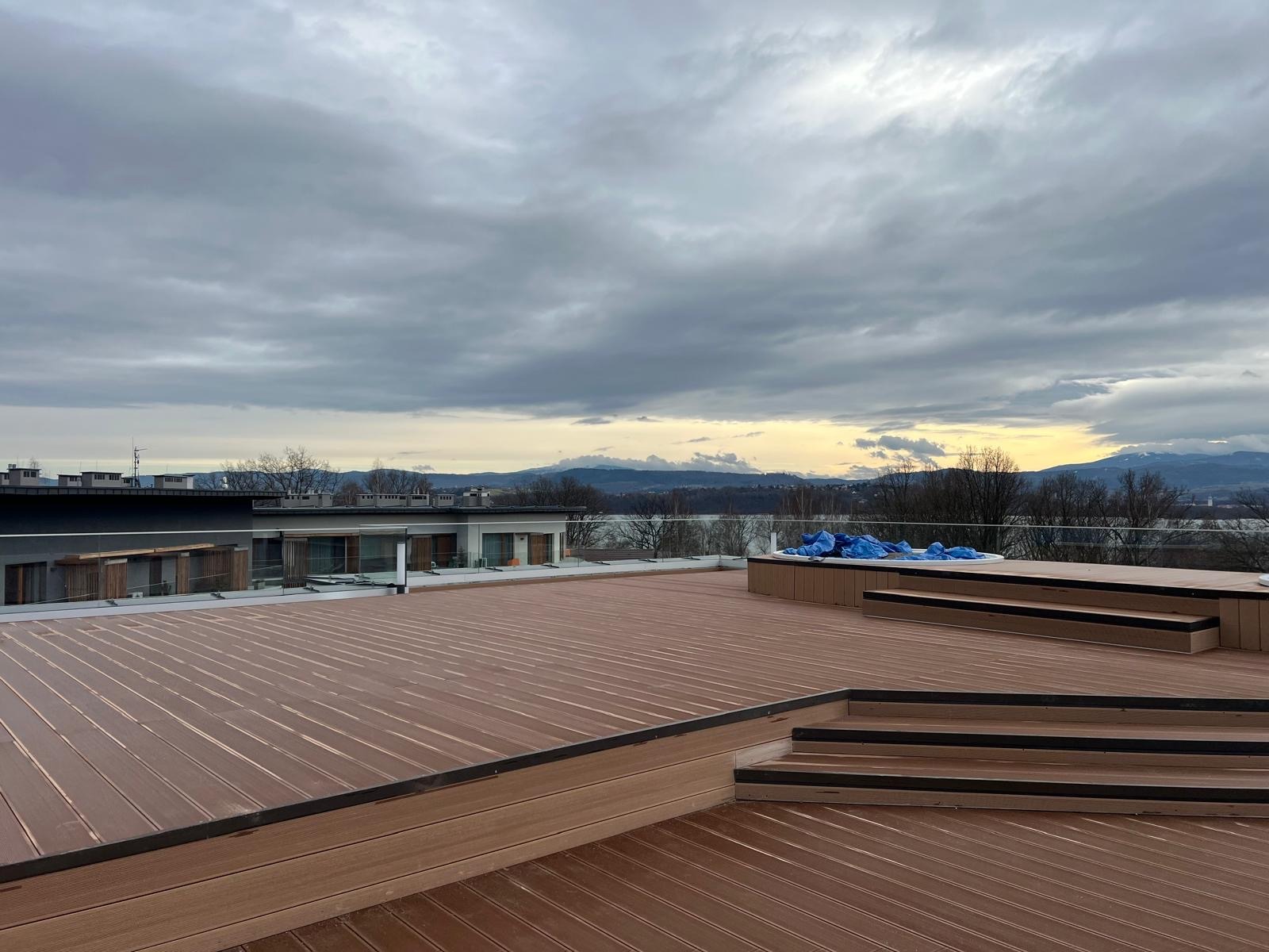 Rozległy taras z brązowych desek, schody z czarnymi akcentami, widok na góry i zachmurzone niebo. W tle budynek z przeszklonymi balustradami i jacuzzi przykryte folią.