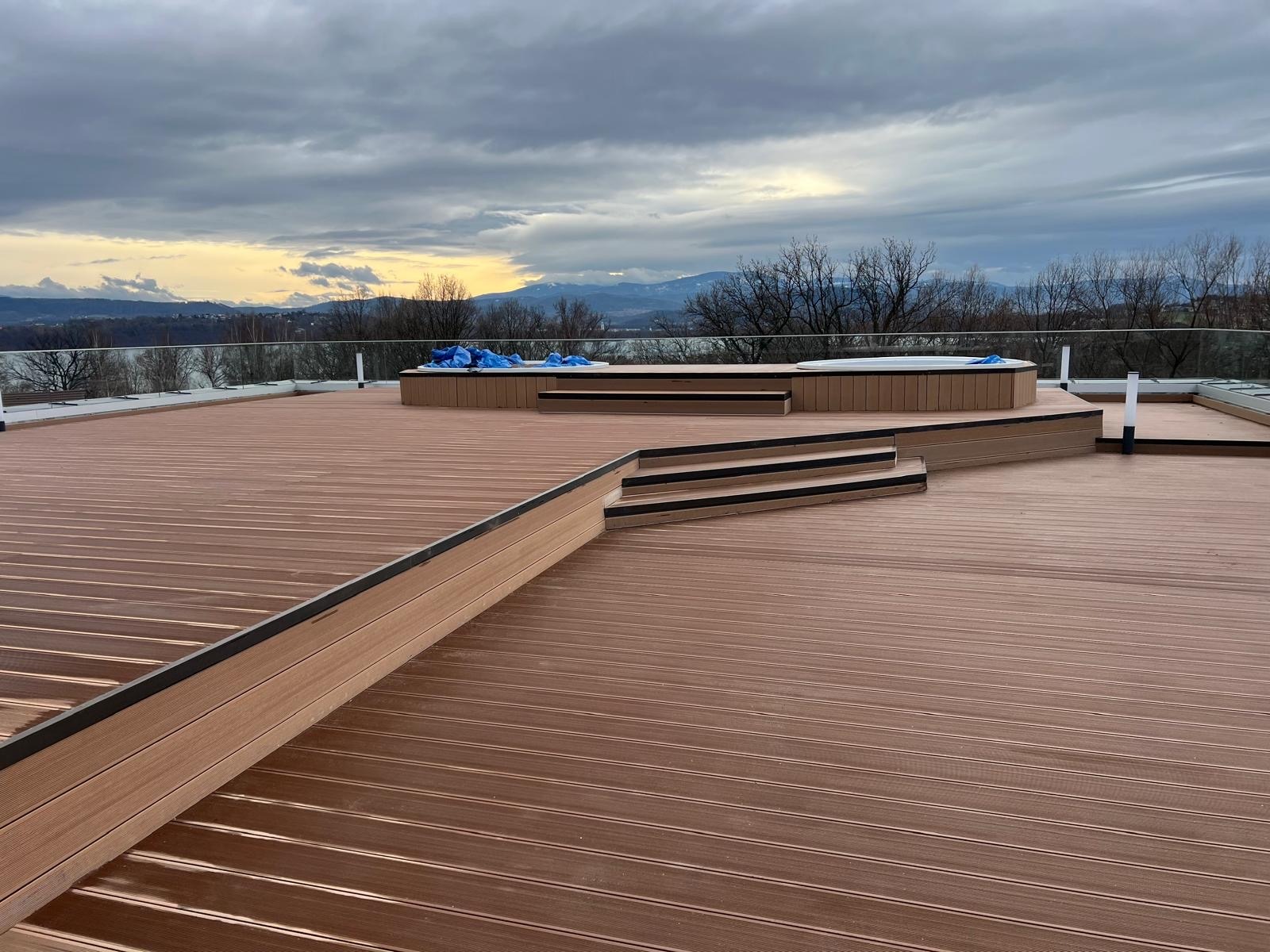 Rozległy taras z brązowych desek kompozytowych z jacuzzi i schodami, otoczony szklaną balustradą, z widokiem na góry i pochmurne niebo. Nowoczesny design.