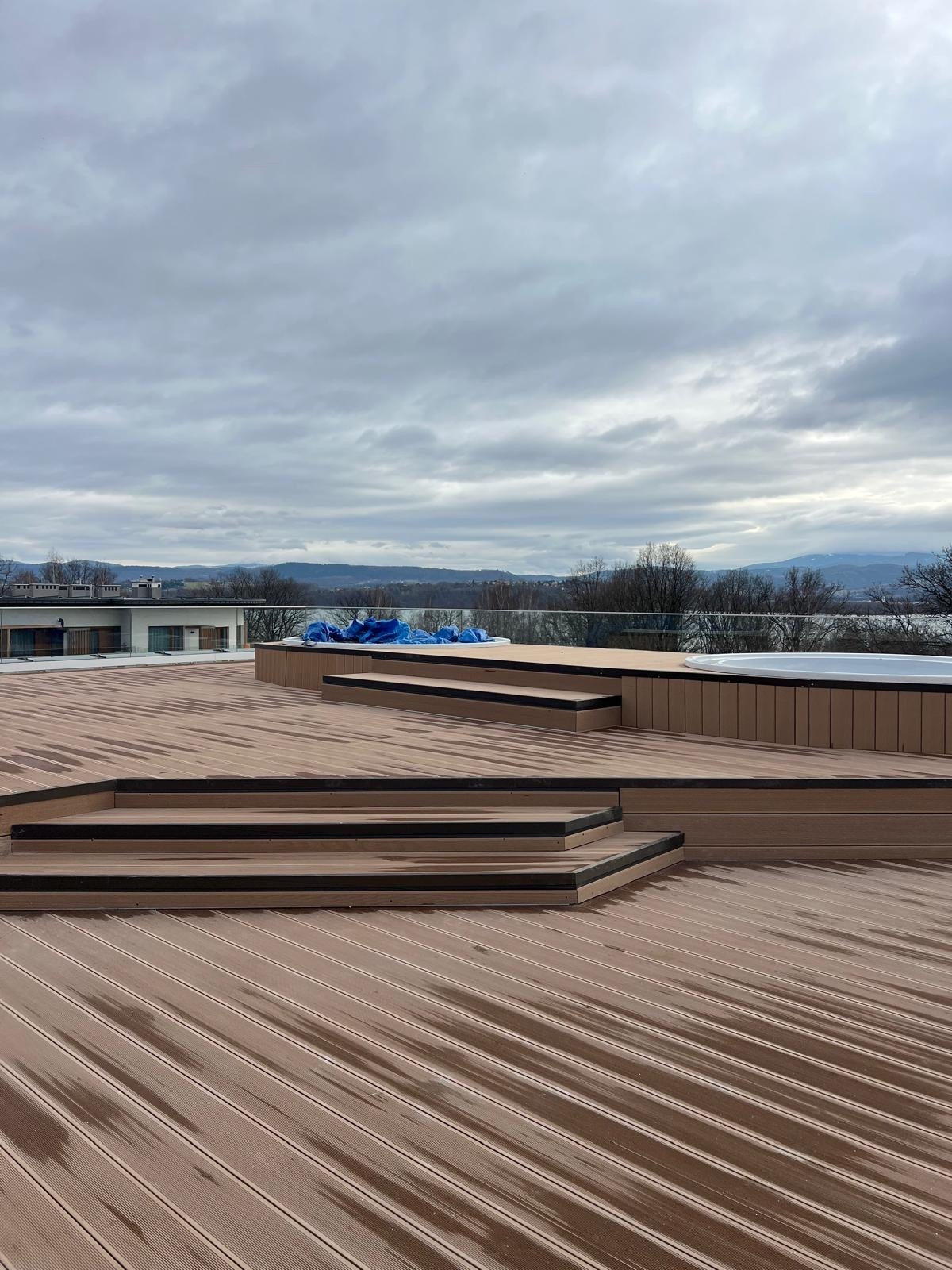 Brązowy taras z kompozytu z schodami i balustradą, widok na jezioro i góry w tle, pochmurne niebo. Część tarasu przykryta folią ochronną.