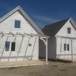 Dwa identyczne, białe domki letniskowe z drewnianą pergolą przed wejściem, ustawione obok siebie na piaszczystym gruncie w słoneczny dzień.