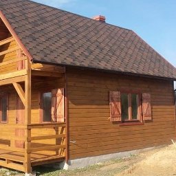 Firma Handlowa MS - Drewniany domek letniskowy z brązowym dachem bitumicznym, drewnianymi okiennicami i małym balkonem, otoczony polną scenerią z widocznym piaskiem i odległym ogrodzeniem.