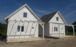 Dwa identyczne, białe domki letniskowe z drewnianą pergolą przed wejściem, ustawione obok siebie na piaszczystym gruncie w słoneczny dzień.