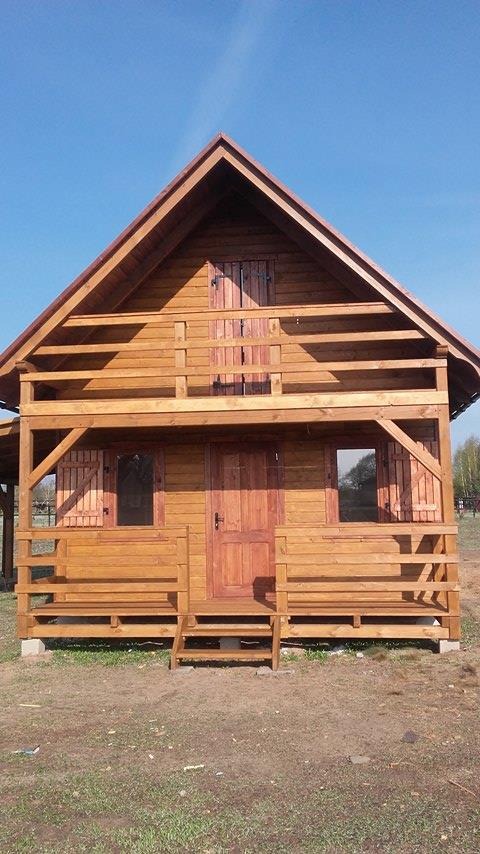 Całkowicie drewniany domek letniskowy z balkonem i schodami wejściowymi, widoczny w pełnym słońcu na tle błękitnego nieba.