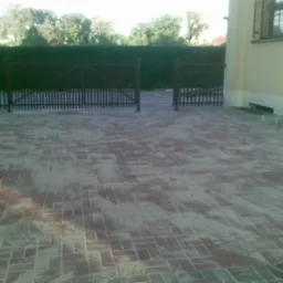 Ułożona kostka brukowa w odcieniach czerwieni i szarości przed posesją z drewnianą bramą i zielonym żywopłotem w tle.