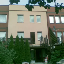 Budynek szeregowy z elewacją w kolorze beżowym i ceglastym, widoczne okna, balkon z drewnianymi schodami, oraz roślinność w postaci krzewów i małych drzewek iglastych.